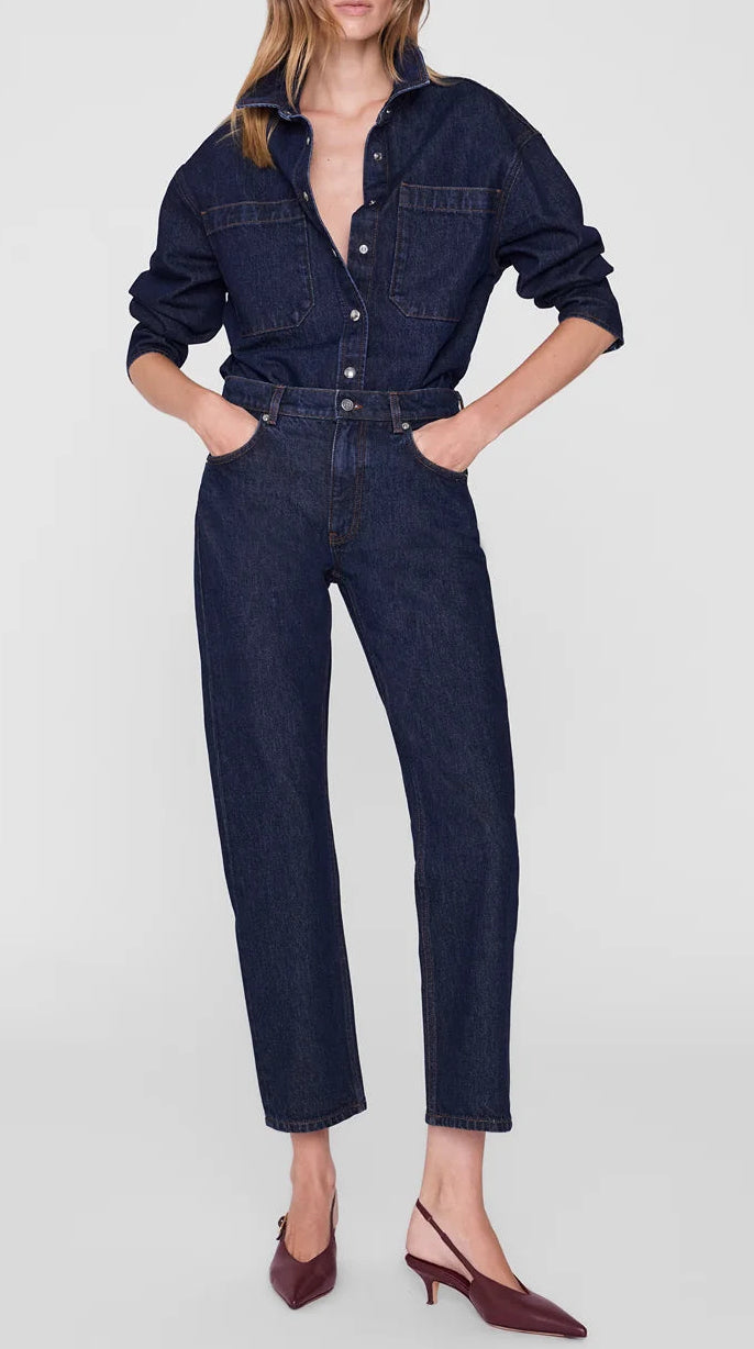 Wearing the Anine Bing Samy Shirt—a dark cotton denim canvas button-up—tucked into matching high-waisted jeans, with hands in pockets and paired with maroon slingback heels.