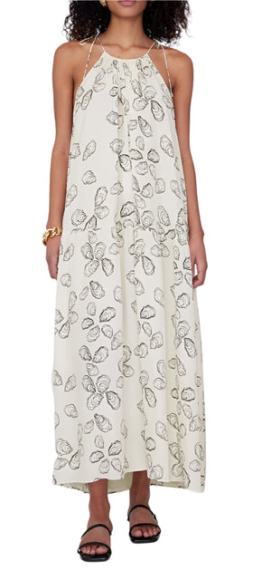 A person is wearing the elegant Santana Dress by Anine Bing, a sleeveless halter-neck maxi with an abstract oyster print in a viscose-silk blend. It's paired beautifully with black sandals and a gold bracelet for a chic, timeless look.
