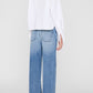 Wearing the Scott Cropped Shirt by Anine Bing, a person stands facing away, styled in a white cotton poplin long-sleeve shirt paired with high-waisted, light blue wide-leg jeans featuring visible back pockets.