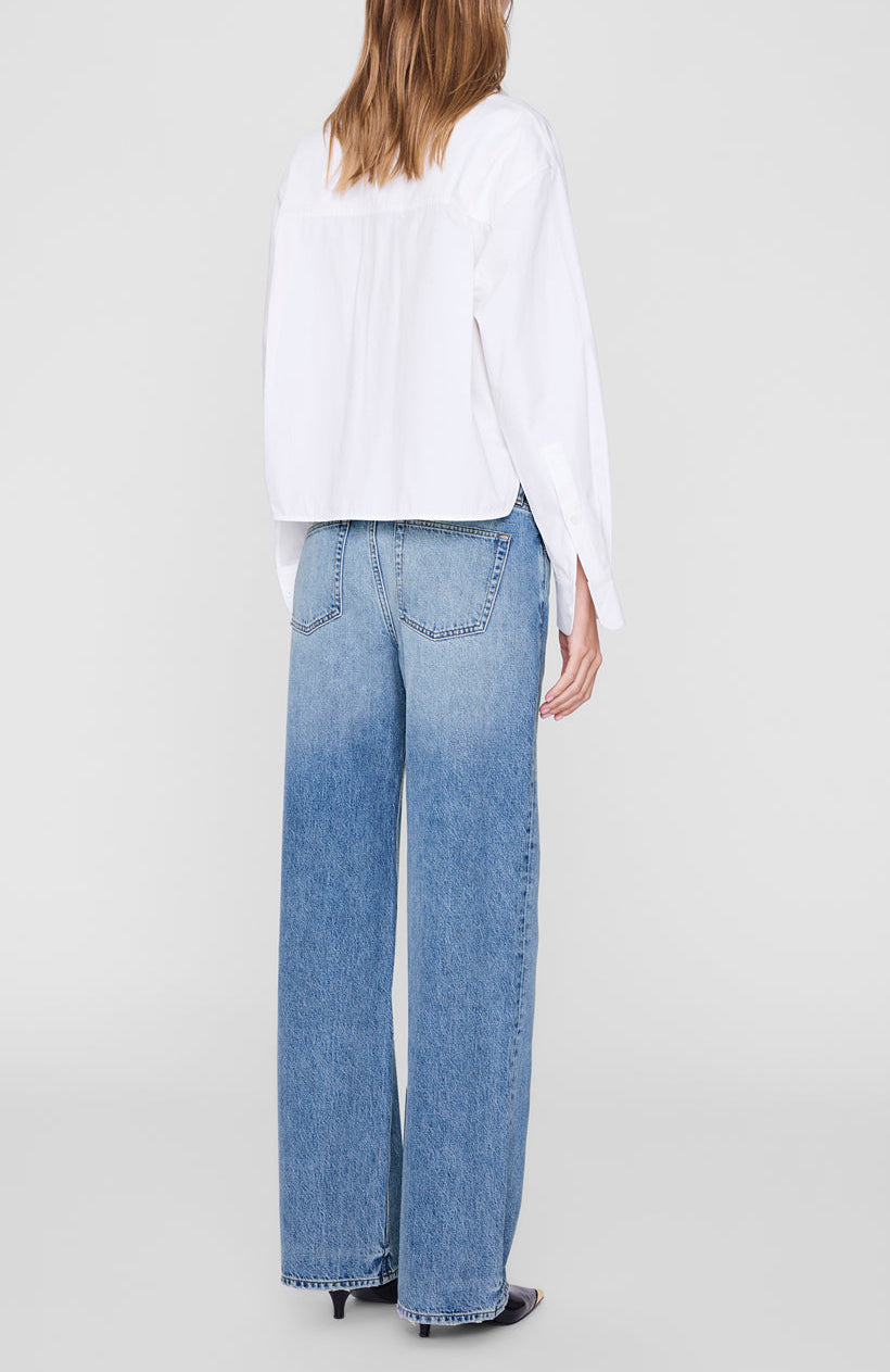 Wearing the Scott Cropped Shirt by Anine Bing, a person stands facing away, styled in a white cotton poplin long-sleeve shirt paired with high-waisted, light blue wide-leg jeans featuring visible back pockets.