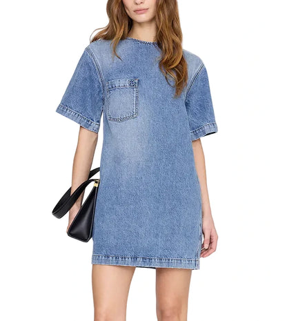 A woman wears the Tulah Dress by Anine Bing—a short-sleeve blue denim mini with a chest pocket—paired with a black handbag, perfect for everyday wear.