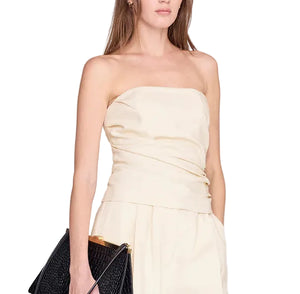 A woman wearing the versatile, strapless Tyla Top by Anine Bing in cream, paired with matching pants, holds a large black textured clutch bag with a sculpted shape.
