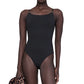 A person wears the Anine Bing Vesta One Piece, a black minimalist swimsuit with adjustable straps, and holds a leopard print garment in one hand against a white background.