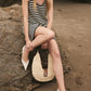 A woman in an Anine Bing Arie Dress with a breezy silhouette sits on a beach rock, holding a woven bag, legs crossed, and wearing white flats.