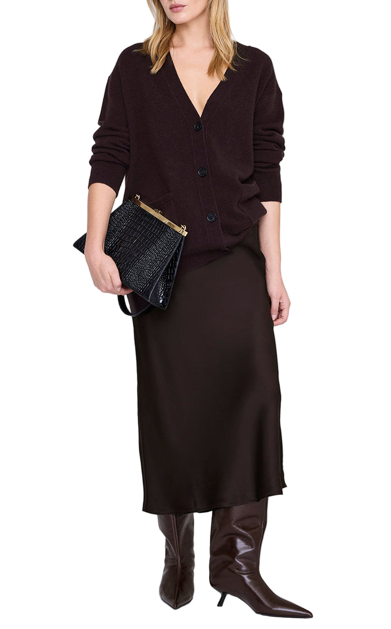 A woman wearing a black cardigan, Anine Bing Bar Silk Skirt, knee-high brown boots, and holding a black textured clutch bag.