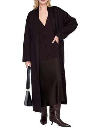 A woman stands on a white background wearing a long black coat, black cardigan, brown knee-high boots, holding a black handbag, and styled with the Anine Bing Bar Silk Skirt.