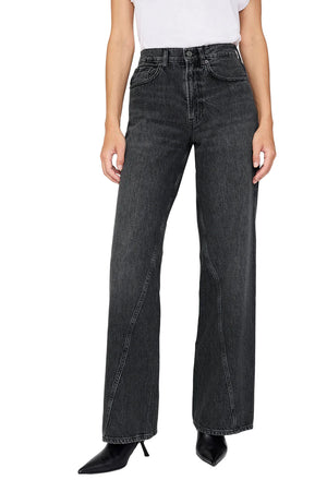 Wearing the Anine Bing Brie Jean—high-waisted, dark wash, wide-leg jeans in rigid cotton denim—paired with a white tee and black pointed-toe boots, standing against a plain background.