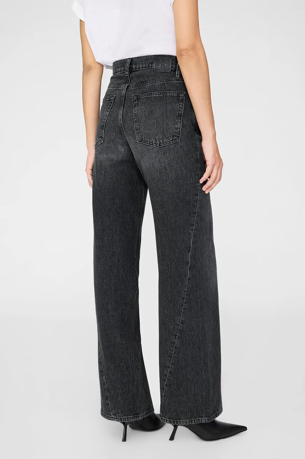 A person is shown from the back wearing Anine Bing's Brie Jean, featuring a high-waisted, wide-leg silhouette in dark wash rigid cotton denim, paired with black heeled shoes against a plain backdrop.
