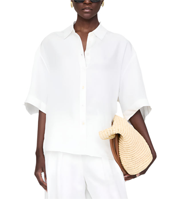 Wearing the Coco Shirt by Anine Bing in ivory and white pants, a person holds a woven straw clutch bag.