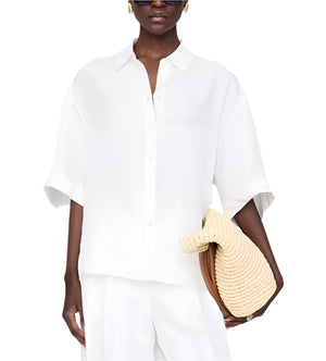 Wearing the Coco Shirt by Anine Bing in ivory and white pants, a person holds a woven straw clutch bag.