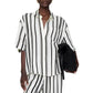 Person wearing the Anine Bing Coco Shirt Monogram Stripe—a boxy collared shirt in a lightweight cupro blend with ivory and black stripes—paired with matching pants and holding a black textured clutch.