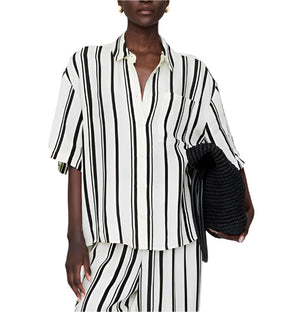 Person wearing the Anine Bing Coco Shirt Monogram Stripe—a boxy collared shirt in a lightweight cupro blend with ivory and black stripes—paired with matching pants and holding a black textured clutch.