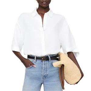 Person wearing the Anine Bing Coco Shirt, a boxy collared ivory top, tucked into light blue jeans, holding a straw hat and brown bag with one hand in their pocket.