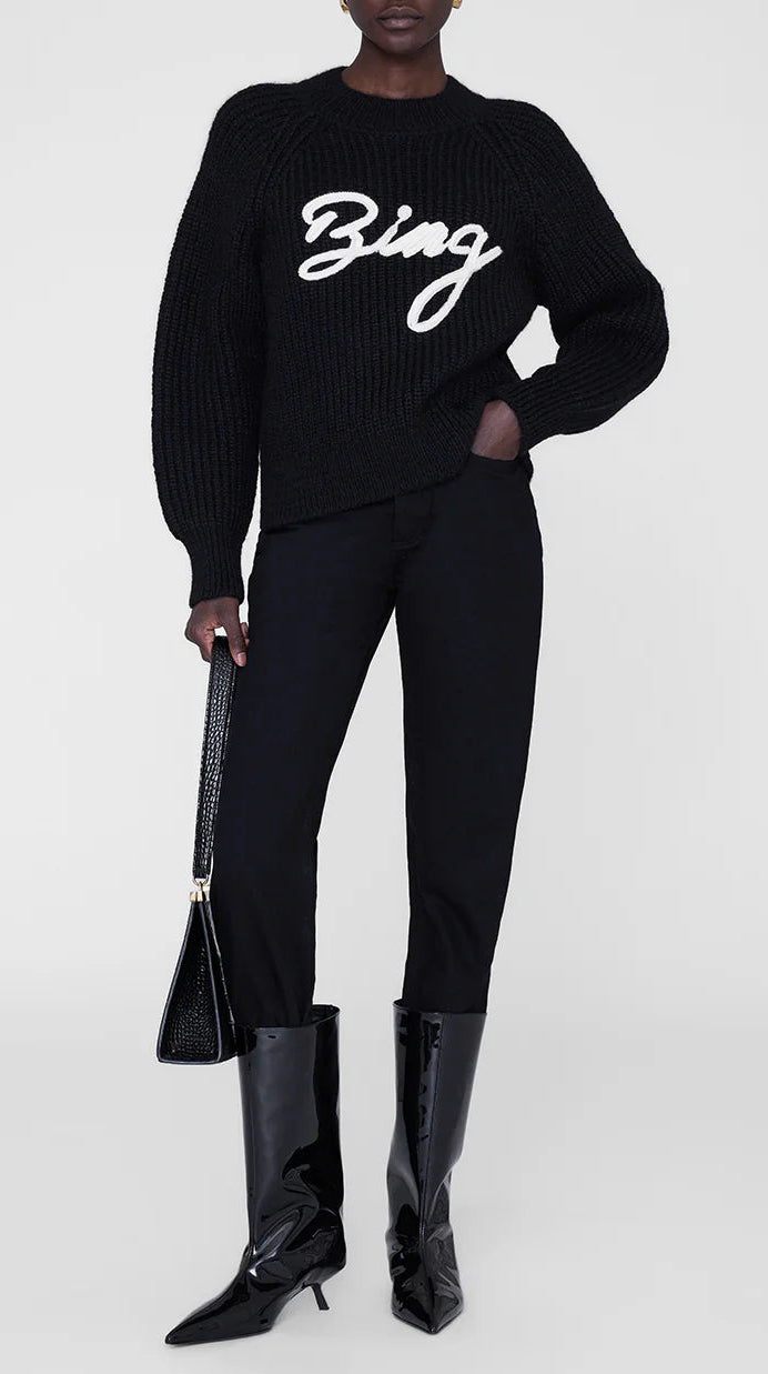 A person in an Anine Bing Kendrick Sweater Bing, black pants, patent boots, and a black handbag stands against a plain background, embodying modern AB branding style.
