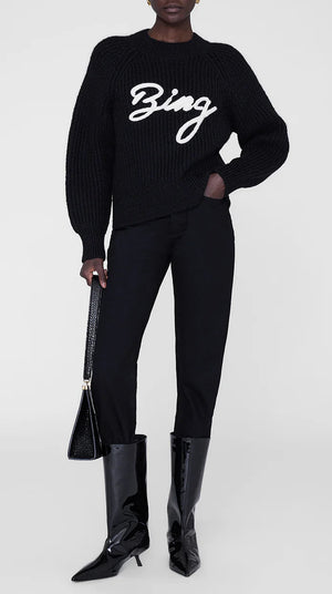 A person in an Anine Bing Kendrick Sweater Bing, black pants, patent boots, and a black handbag stands against a plain background, embodying modern AB branding style.