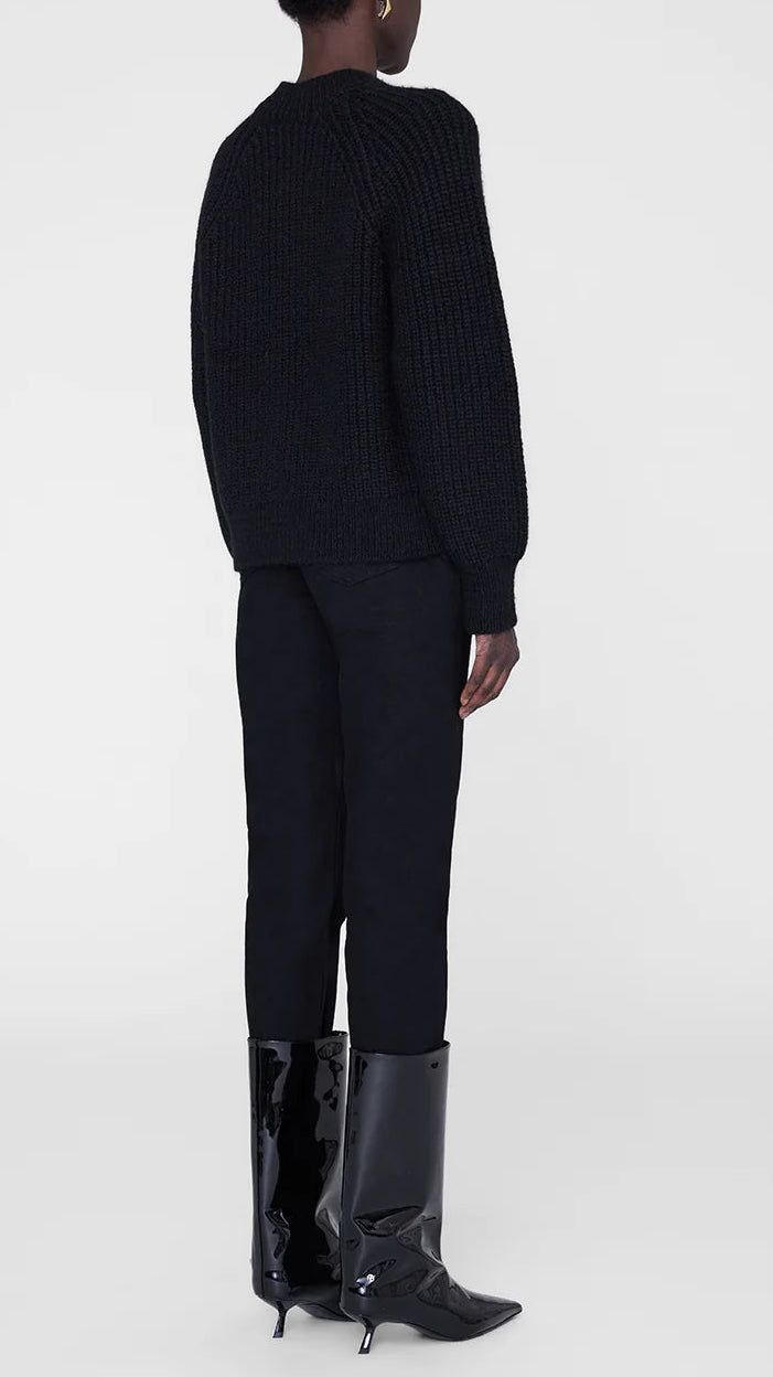 Person standing against a plain background, wearing a black Kendrick Sweater Bing by Anine Bing, black pants, and black patent knee-high heeled boots—an elevated look with a subtle nod to AB branding.