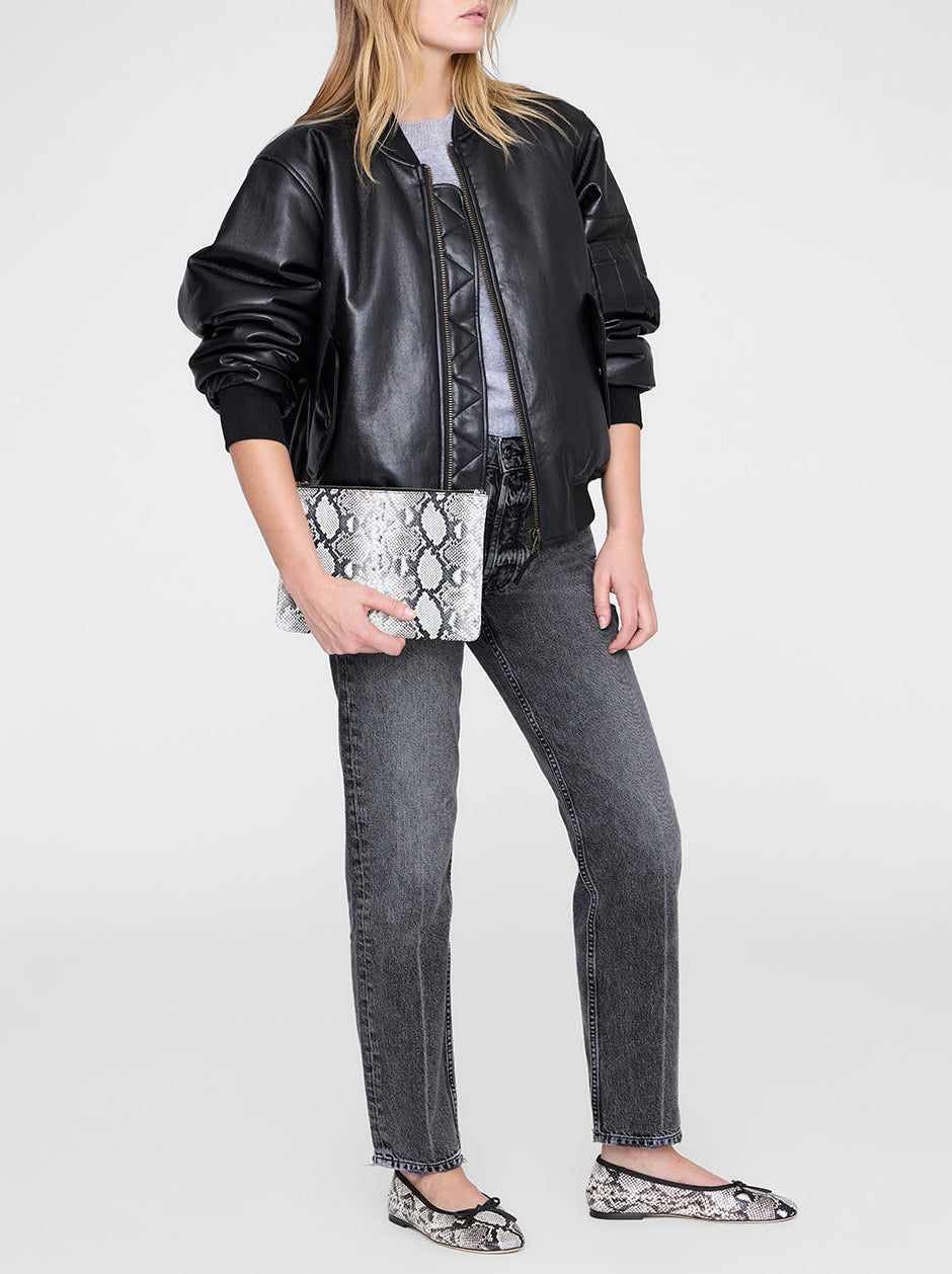 A woman wearing a black leather jacket, gray shirt, gray jeans, and snakeskin-patterned flats holds the Anine Bing Elly Large Pouch with a chic python print against a plain background.