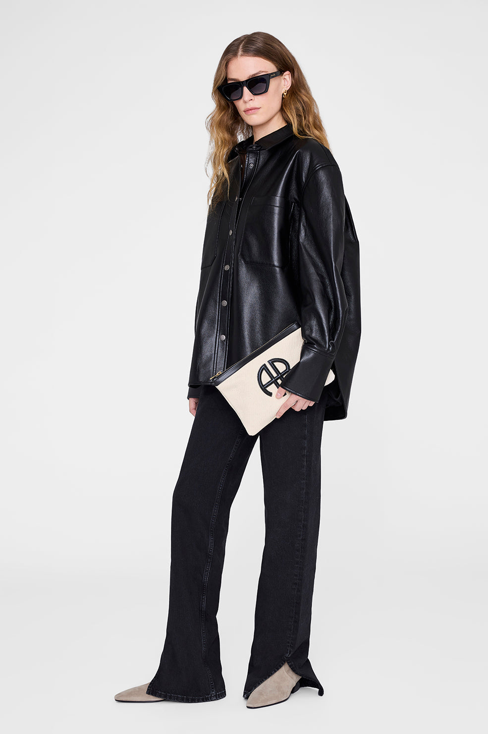 A woman in black sunglasses, a black leather shirt, black jeans, and beige pointed shoes holds an Anine Bing Lili Canvas Pouch with a black logo, standing against a plain white background.