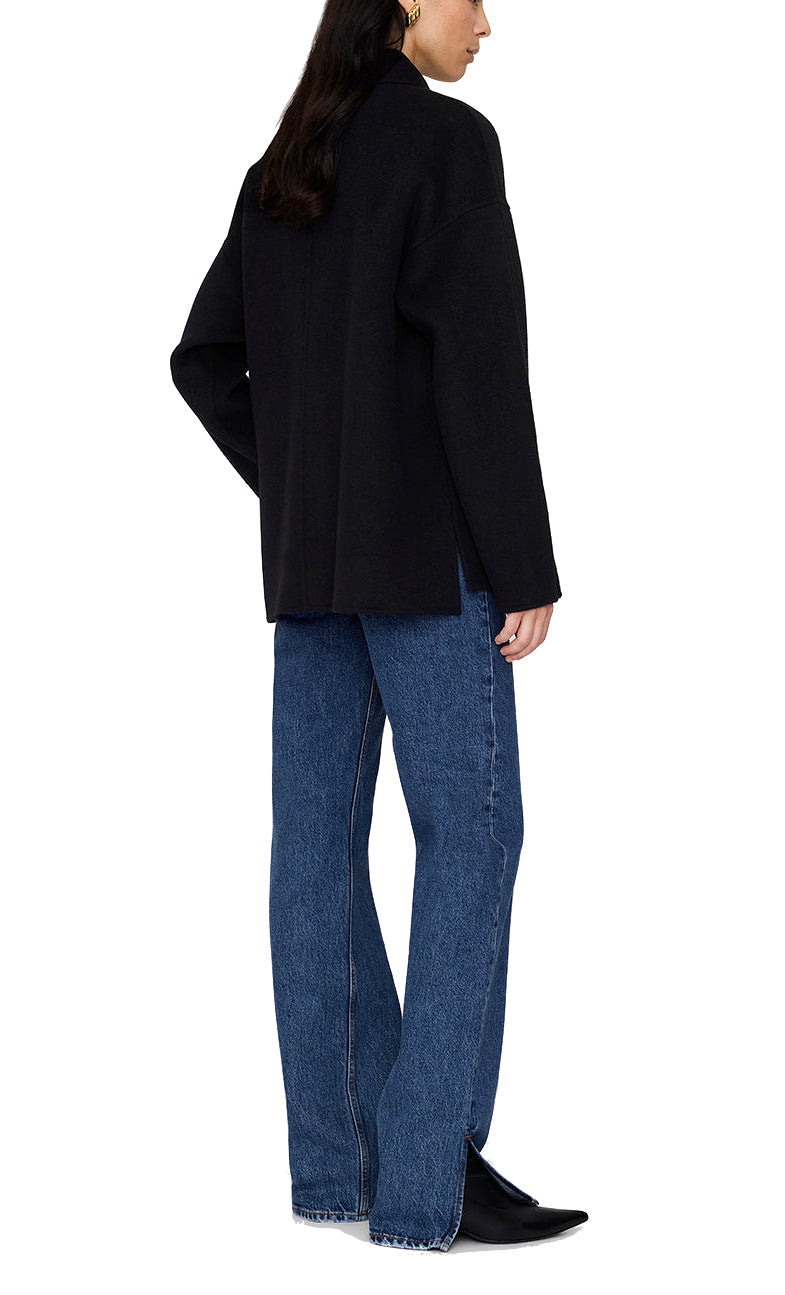 Someone stands at a slight angle facing away, wearing an Anine Bing Luca Cashmere Blend Jacket in black, paired with blue straight-leg jeans and black pointed-toe shoes.