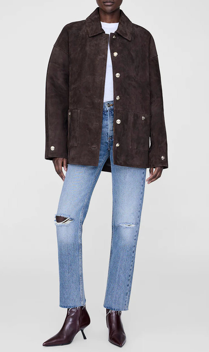 A person in the Anine Bing Luca Suede Jacket—a dark brown button-up—paired with a white shirt, ripped light blue jeans, and pointed dark brown ankle boots stands against a plain background.