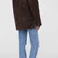 A person stands with their back to the camera, wearing the Anine Bing Luca Suede Jacket in dark brown, paired with light blue jeans and pointed maroon ankle boots.