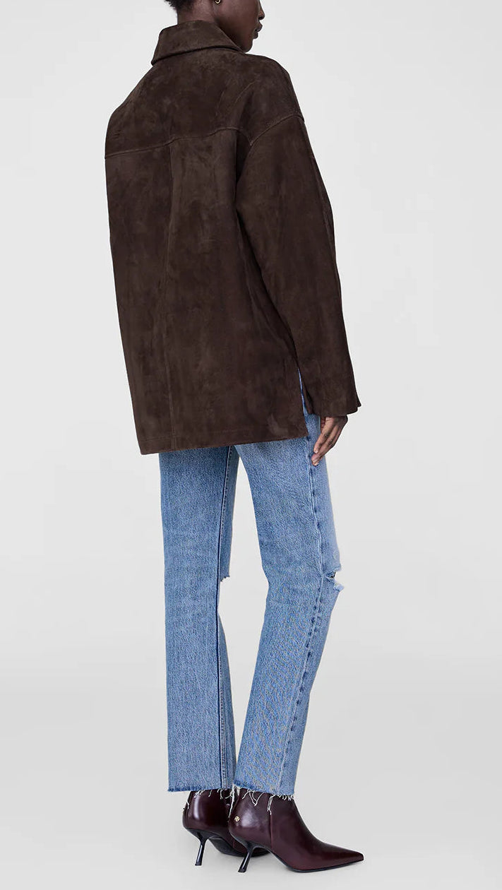 A person stands with their back to the camera, wearing the Anine Bing Luca Suede Jacket in dark brown, paired with light blue jeans and pointed maroon ankle boots.