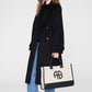 A person in a long black coat, blue jeans, and black boots holds the Anine Bing Medium Lara Tote—a beige and black cotton canvas bag with leather accents and "AB" initials. They have long hair and sunglasses.