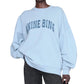 A person wearing the ANB Miles Sweatshirt by Anine Bing—a pale blue oversized cotton fleece crewneck with "ANINE BING" on the front—paired with dark pants and holding a black bag.