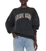 A person wearing the Anine Bing Miles Leopard Sweatshirt—an oversized black style crafted from organic cotton fleece with leopard print lettering—paired with light blue jeans, stands with one hand in their pocket.
