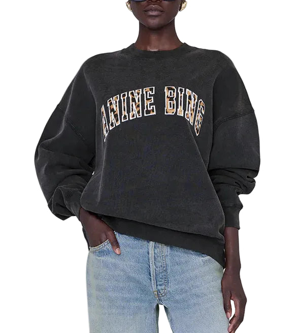 A person wearing the Anine Bing Miles Leopard Sweatshirt—an oversized black style crafted from organic cotton fleece with leopard print lettering—paired with light blue jeans, stands with one hand in their pocket.