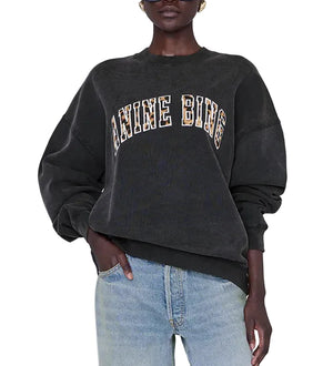 A person wearing the Anine Bing Miles Leopard Sweatshirt—an oversized black style crafted from organic cotton fleece with leopard print lettering—paired with light blue jeans, stands with one hand in their pocket.
