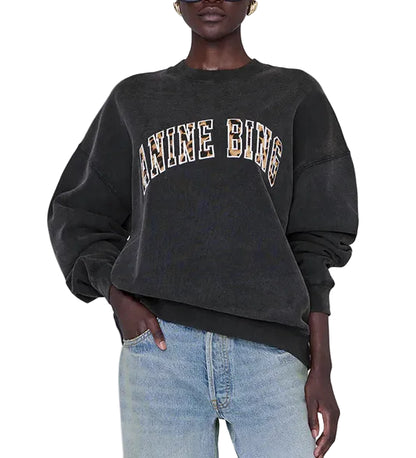 A person wearing the Anine Bing Miles Leopard Sweatshirt—an oversized black style crafted from organic cotton fleece with leopard print lettering—paired with light blue jeans, stands with one hand in their pocket.