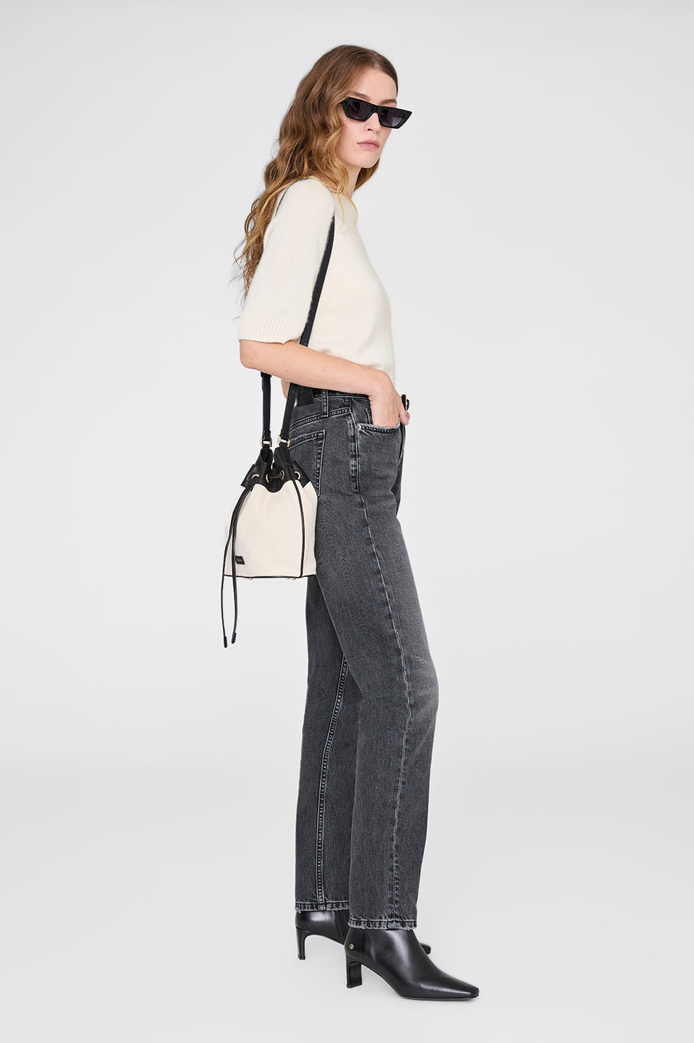 Woman in profile against a plain background wears sunglasses, a cream short-sleeve sweater, gray jeans, black ankle boots, and carries the Anine Bing Mini Alana Bucket Bag in cream and black with gold-tone hardware.