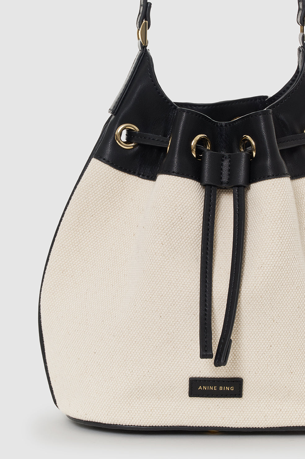 The Mini Alana Bucket Bag by Anine Bing features beige canvas, black leather trim, gold-tone hardware, a drawstring closure, adjustable strap, and "Anine Bing" label on the front.