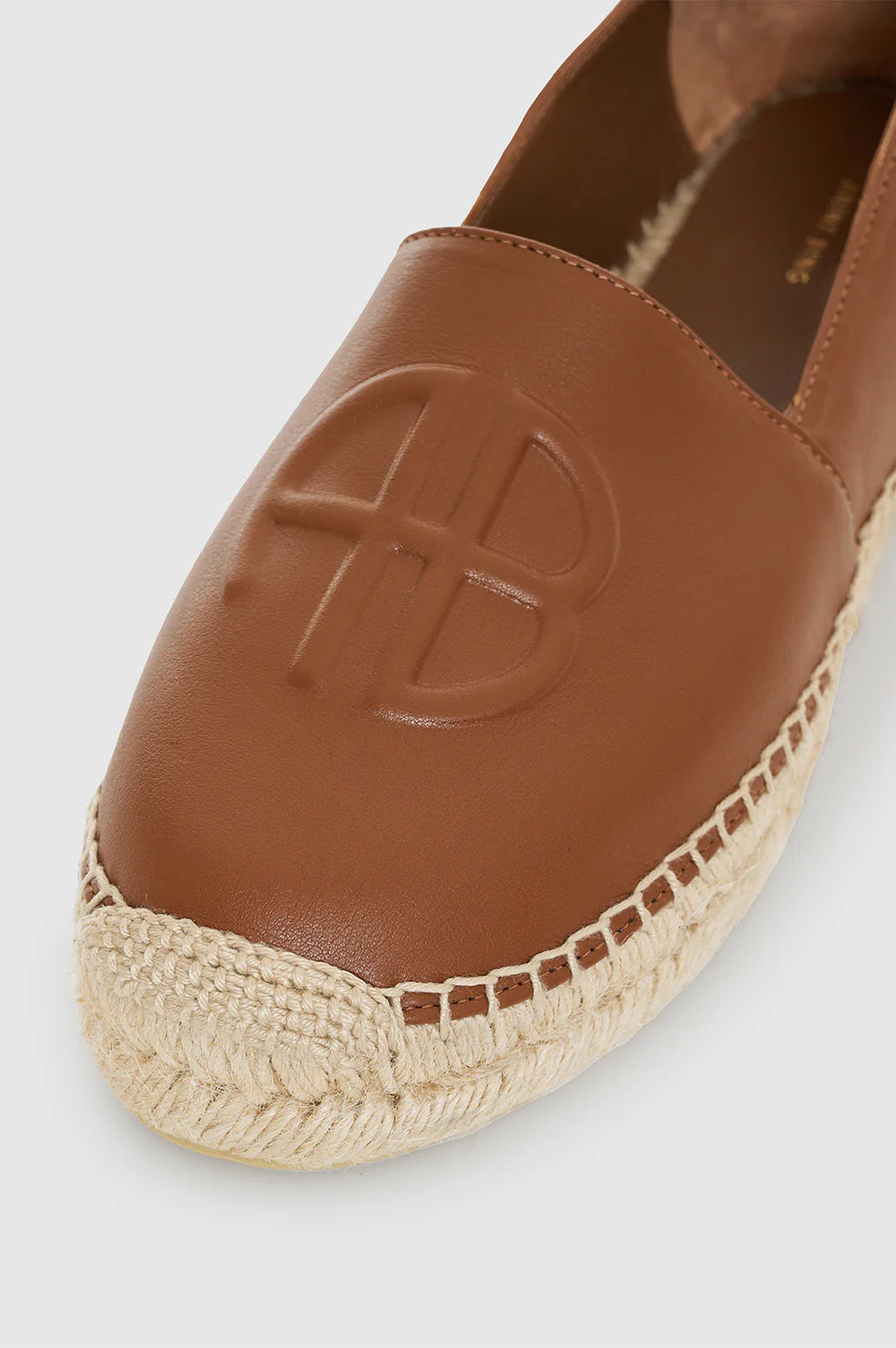 Close-up of the Paloma Espadrilles by Anine Bing, featuring a brown leather upper embossed with "AB" initials and a woven jute sole.