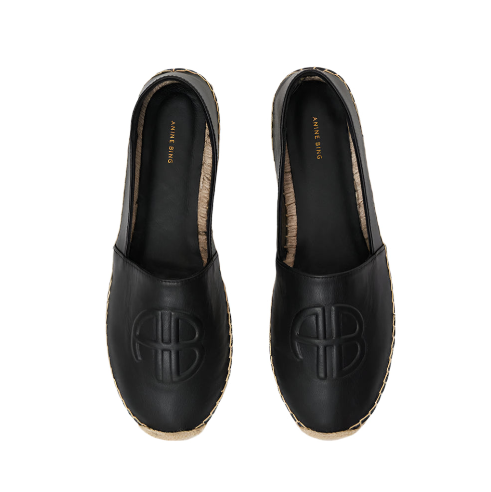 The Paloma Espadrilles by Anine Bing are black leather espadrilles with a woven sole and front embossing, featuring the gold ANINE BING monogram on the insole.