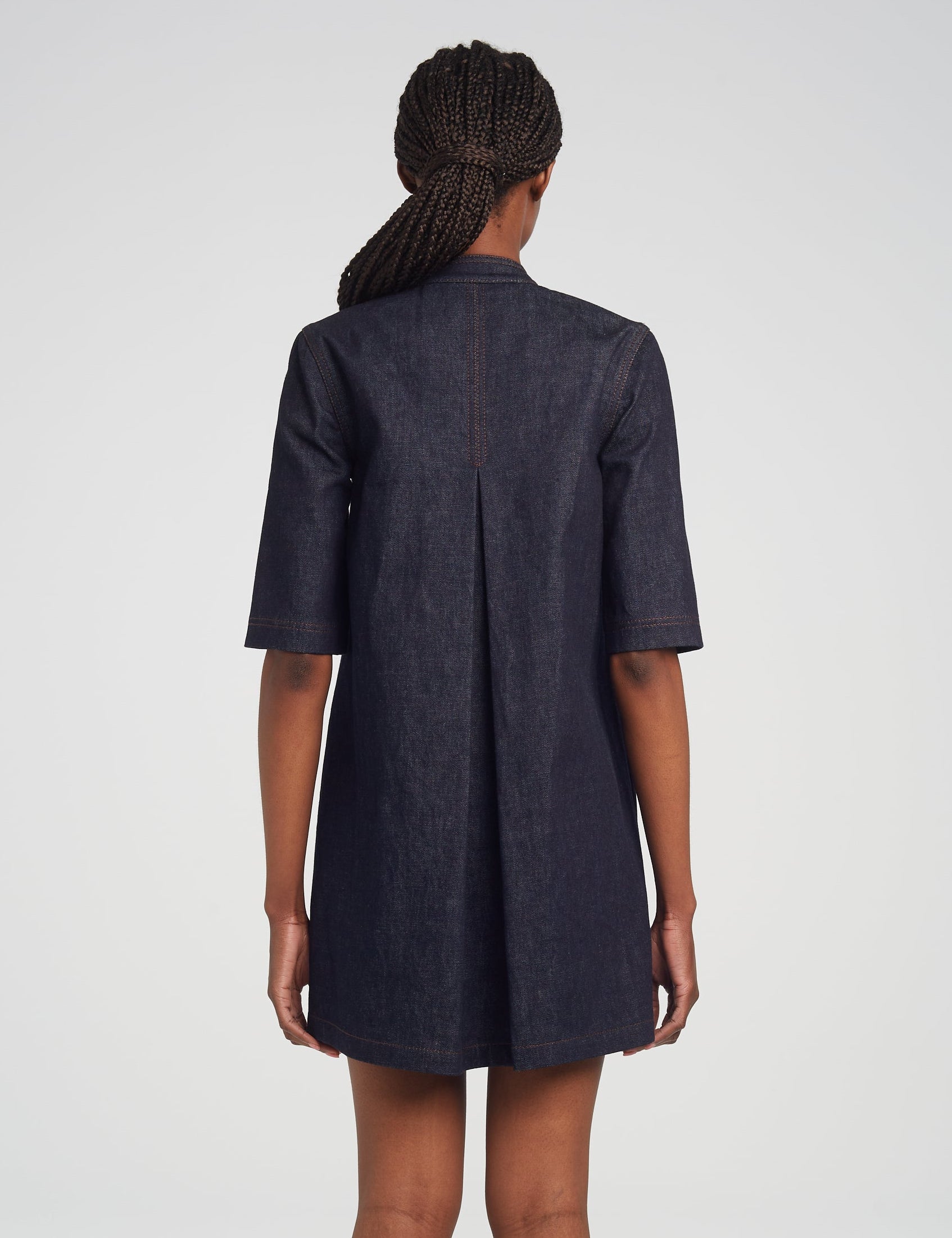 A person with braided hair, seen from behind, is wearing the Adam Lippes Lauder Dress—a dark Japanese indigo denim short-sleeve dress featuring a center back pleat and subtle topstitching.