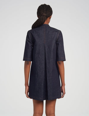 A person with braided hair, seen from behind, is wearing the Adam Lippes Lauder Dress—a dark Japanese indigo denim short-sleeve dress featuring a center back pleat and subtle topstitching.