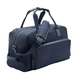 The Peter Millar Pursuit Duffle is a navy blue travel bag featuring black zippers, handles, an adjustable shoulder strap, and a front laptop zipper pocket, shown on a white background.