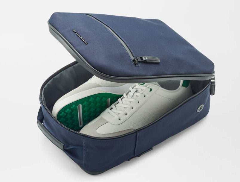 The Peter Millar Pursuit Shoe Bag in navy, featuring genuine leather trim and a zippered closure, is shown partially open to reveal white sneakers with green accents against a white background.