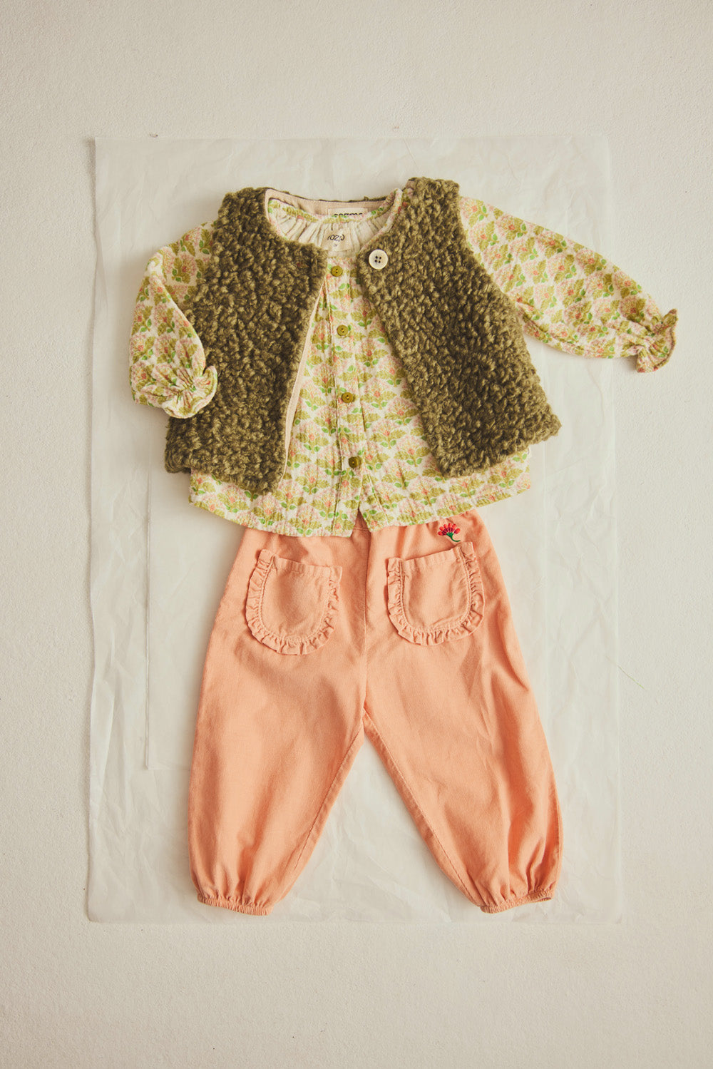 A baby outfit featuring a patterned long-sleeve top, a textured green vest, and Cozmo Baby Girls’ Agen Corduroy Pants in peach with ruffled pockets, laid flat on a white background.