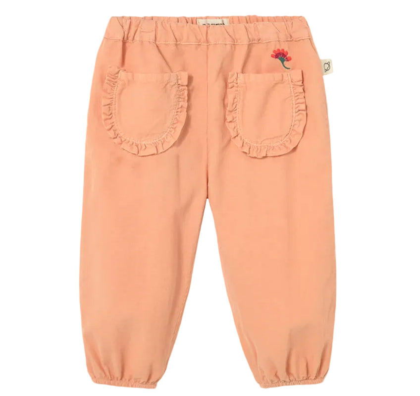 The Cozmo Baby Girls’ Agen Corduroy Pants feature a peach-colored micro-corduroy fabric, an elastic waist and cuffs, two ruffled back pockets, and a small embroidered red flower above the right pocket.