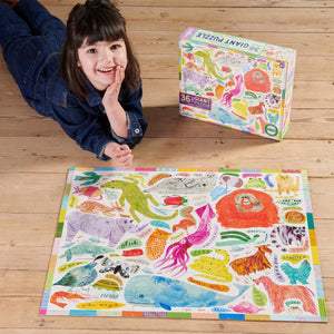A child lies on a wooden floor next to a completed, colorful Eeboo Animal Voices 36 Piece Giant Puzzle, its box labeled "36 Giant Puzzle.