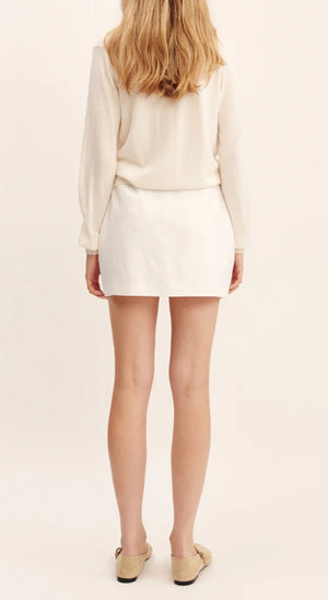 A person stands facing away, wearing a long-sleeved cream top, beige shoes, and the white linen Alice Skirt with an A-line silhouette by POSSE.