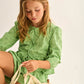 A young girl wearing the Cozmo Girls' Alma Eyelet Blouse sits on a stool and looks down at a mesh bag containing yellow fruits against a plain background.