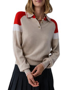 A woman wears the Guest in Residence Alpine Collegiate Polo, a beige women’s cashmere sweater with red and white shoulder accents, paired with a pleated black skirt against a white background.