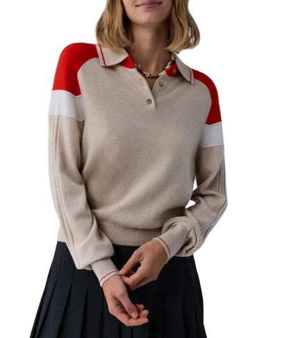 A woman wears the Guest in Residence Alpine Collegiate Polo, a beige women’s cashmere sweater with red and white shoulder accents, paired with a pleated black skirt against a white background.
