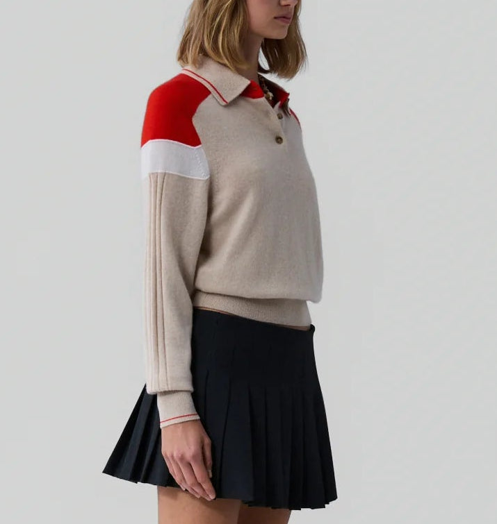 A woman wears the Guest in Residence Alpine Collegiate Polo, featuring red and white shoulder accents, paired with a pleated black skirt, standing against a plain light gray background.