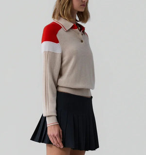 A woman wears the Guest in Residence Alpine Collegiate Polo, featuring red and white shoulder accents, paired with a pleated black skirt, standing against a plain light gray background.