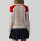 A person stands facing away, wearing the Guest in Residence Alpine Collegiate Polo—a women's cashmere sweater in beige with red and white accents—and a black pleated skirt, set against a plain light gray background.